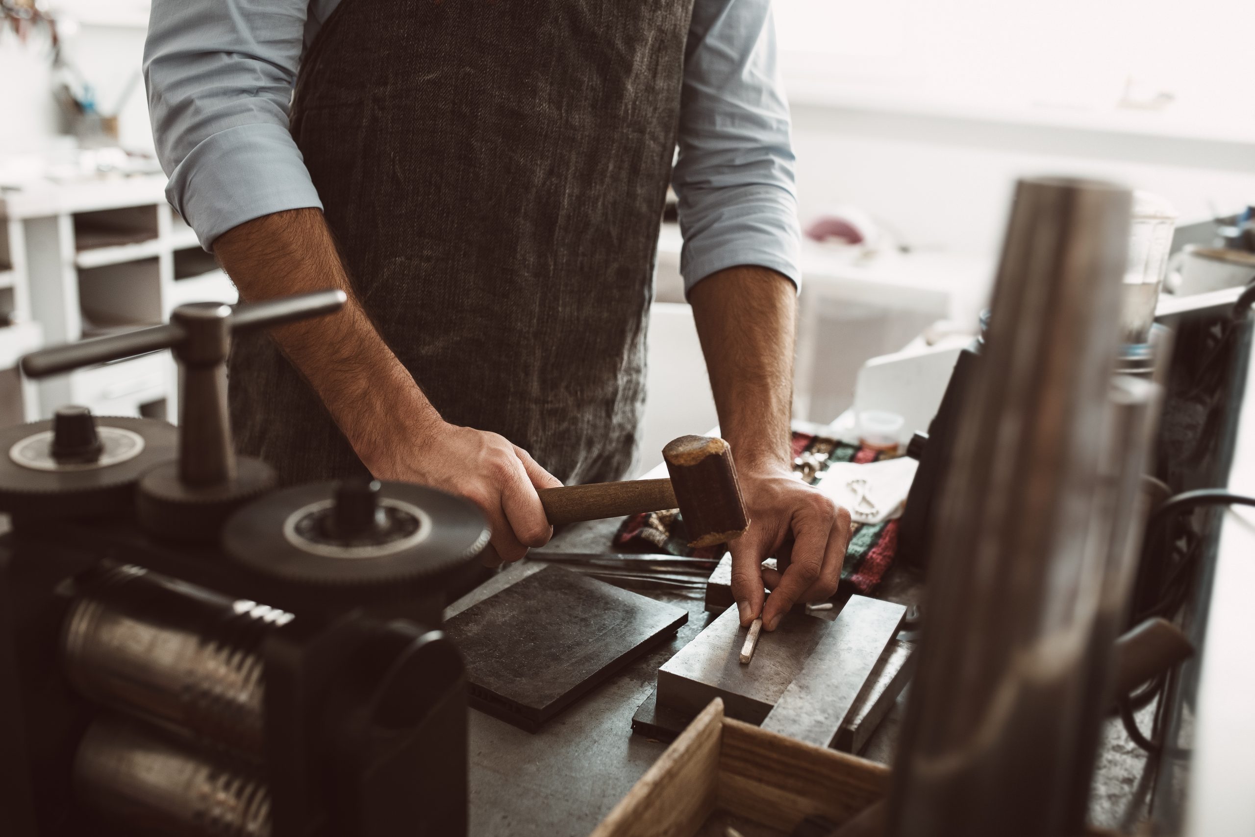Important stage in the work. Male goldsmith wearing apron making a new product at a workbench using hammer